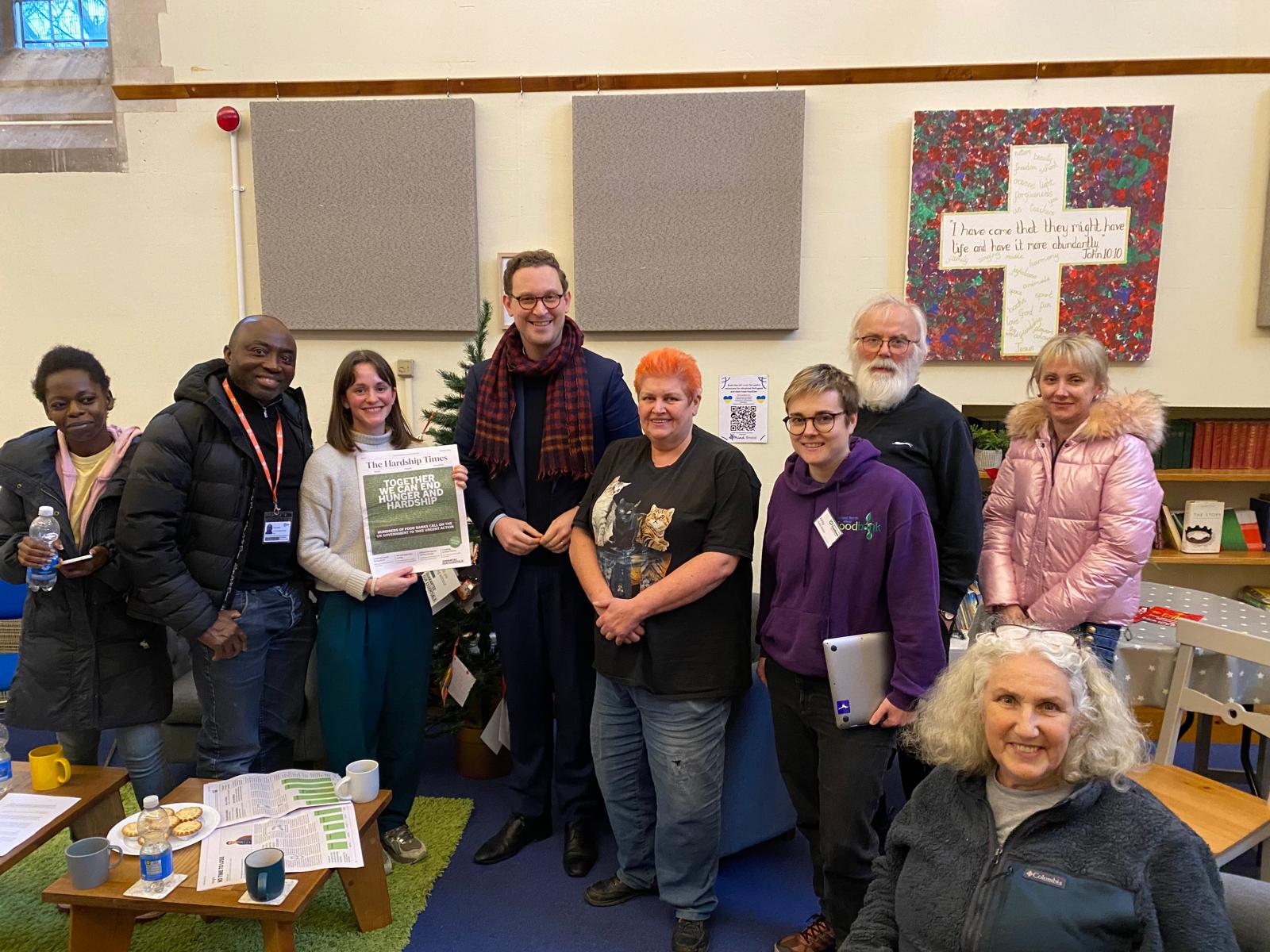 A group photo of community members and staff at Bristol North West Food Bank, standing in front of a decorated Christmas tree and religious artwork. One person holds up a copy of "The Hardship Times" newsletter. The room features bookshelves and acoustic panels.